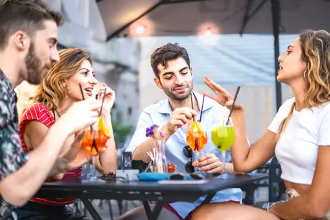 Four trendy young people get together carefree sitting and bonding at outdoors bar restaurant - best friends having fun drinking cocktails at summer a